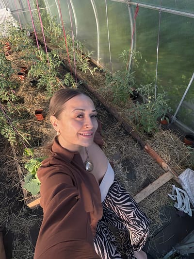 In a greenhouse in Kansk, Russia, a woman with brown hair pulled back in a ponytail poses for a selfie. She is wearing a brown open-collar shirt, a white top, and black and white zebra-print pants. Her left arm is extended, holding the camera. Her right hand is visible near her lap. A large, round, silver pendant hangs from a chain around her neck. The greenhouse is filled with rows of tomato plants, supported by stakes and strings. The ground is covered in straw. Wooden planks form the raised beds for the plants. The greenhouse structure is visible in the background, with translucent panels allowing light to enter.