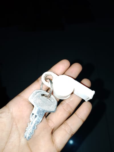A person's hand in Sendangrejo, Indonesia, holds a metal key and a white plastic whistle on a keyring. The background is dark and out of focus, suggesting an indoor or nighttime setting. The key is silver and appears to be a standard house or car key, with serrated edges and a hole for the keyring. The whistle is small and white, with a mouthpiece extending from a bulbous end. The hand is cupped, with fingers gently curled around the objects. The image is a close-up shot, emphasizing the texture of the skin and the details of the objects.