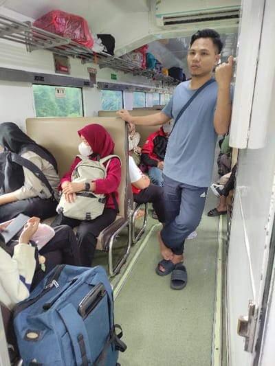 The vertically oriented image captures a crowded interior of a passenger train car, likely in Indonesia given the context of Sukorejo. The setting appears to be an economy or non-reserved class coach, characterized by close quarters and passengers resting in various positions.

In the foreground, a man in a light blue t-shirt and dark pants stands, looking towards the camera. He wears black sandals and has one hand placed on a support beam or seatback. Another person's leg and arm are visible below him, suggesting they are seated or lying on the floor. Further into the carriage, a woman wearing a red hijab and a white face mask is seated, her head slightly down, possibly resting. A white backpack is slung over her shoulder. Other passengers are visible in the background, some lying down in the aisle or between seats, with luggage bags scattered around them.

Notable objects include metal overhead luggage racks, some holding belongings like a red plastic-wrapped bag and other fabric bags. A large blue luggage bag is prominent on the floor in the lower right. Windows line the side of the train, revealing a blurred green landscape outside, indicative of movement through a natural environment. The train interior has light-colored walls and a light green floor. Air conditioning vents are visible on the ceiling.

The scene depicts people traveling and resting during their journey. The time of day appears to be daytime, as natural light filters through the windows. There is no discernible text visible on signs or objects within the image. The overall atmosphere suggests a busy journey.