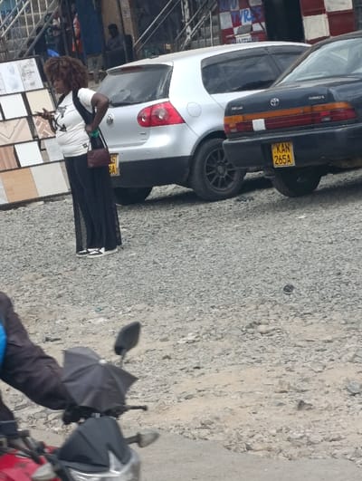 This image depicts an outdoor urban scene in Matungulu west ward, Kenya. The ground is unpaved, composed of light-colored gravel and dirt.
In the mid-ground, a woman stands facing left, holding a black smartphone in her right hand. She has dark, curly hair and is wearing a white t-shirt with a dark graphic, black wide-leg trousers, and white athletic shoes. A small, dark red handbag with a strap is slung over her left shoulder.
Behind the woman, two vehicles are parked on the gravel. The vehicle on the left is a silver Volkswagen Golf hatchback, visible from the rear-left, with a partially obscured Kenyan license plate showing "KG 8A". The vehicle on the right is a dark blue or black Toyota Corolla sedan, visible from the rear-right, displaying the Kenyan license plate "KAN 265A" and featuring a white horizontal stripe across the trunk lid above the license plate.
In the background, to the left, a vertical display rack exhibits multiple rectangular tiles of various patterns and colors. Further back, structures consistent with commercial buildings are visible, including one storefront with the partial word "TYRES" discernible, and metal grates. A dark-clad figure is partially visible further in the background.
In the lower foreground, on the left, the front section of a red and black motorcycle is partially visible, including its handlebars, a rearview mirror, a headlight, and a small black umbrella attached to the handlebars.