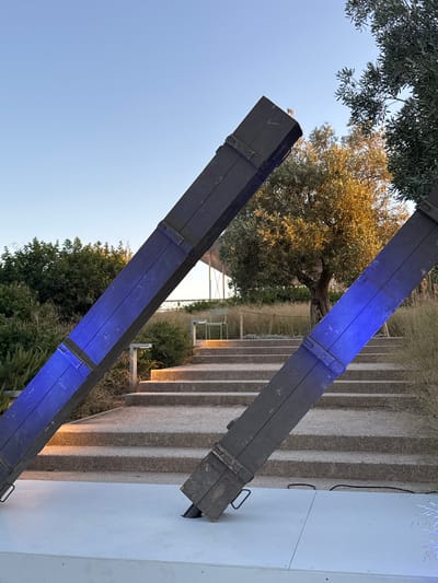 This image depicts an outdoor scene with two large, dark wooden beams angled upwards from a white platform. The beams are illuminated with vibrant blue light, and appear to be part of an art installation or architectural feature. Behind the beams, a set of wide, stone stairs ascends a gentle slope, flanked by lush green foliage and dry, wispy grasses. An olive tree with golden leaves stands prominently in the mid-ground, catching the sunlight. In the background, a modern structure with white supports and a translucent roof is visible, suggesting a public space like a park or an outdoor venue. The sky is clear and bright blue, indicating it is daytime. The scene is set in the Municipal Unit of Dafni, Greece. There are no people visible in the image.