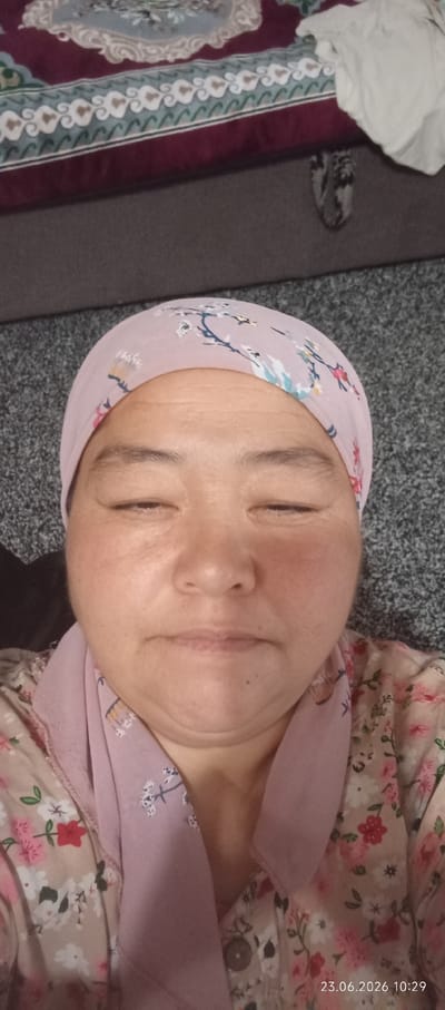A frontal close-up photo of an adult woman wearing a light pink floral headscarf and a matching floral shirt is presented. Her eyes are closed, and her head is tilted slightly downwards. The background features a grey carpeted surface and a patterned fabric, possibly a blanket or tapestry, in the upper portion of the frame. A timestamp "23.06.2026 10:29" is visible in the lower right corner of the image. The photograph was taken indoors, as indicated by the presence of soft furnishings and the lighting. The specific location within the city of Chon-Kara, Kyrgyzstan, cannot be determined from the visual information provided.