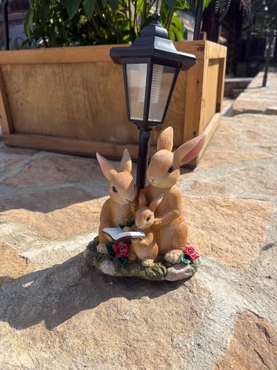 A solar-powered lantern figurine stands on a stone pathway in what appears to be a residential garden. The figurine depicts a family of three brown rabbits gathered around an open book, with two red roses at the base. The larger rabbits appear to be reading to the smallest one. Behind the figurine is a wooden planter box filled with green plants. The setting sun casts long shadows on the stone pathway. The image does not contain any information that would suggest the location is in Колюткино, Russia.