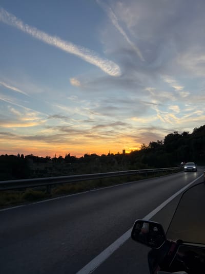 This is a photograph taken at dusk from a vehicle traveling on a road in Gornji Katun, Montenegro. The sky is painted with the warm hues of sunset, with oranges, yellows, and blues blended with scattered wispy clouds. In the distance, silhouetted against the vibrant sky, are the shapes of trees and buildings, including what appears to be a church steeple and possibly a tower. The road ahead is paved, with a white line marking the lane. A metal guardrail separates the road from the embankment. Several cars are visible on the road, moving in the same direction. In the foreground, a side-view mirror of the vehicle is visible, reflecting a passenger. The surrounding landscape is hilly and covered with dense, dark green foliage, suggesting a rural or semi-rural setting.