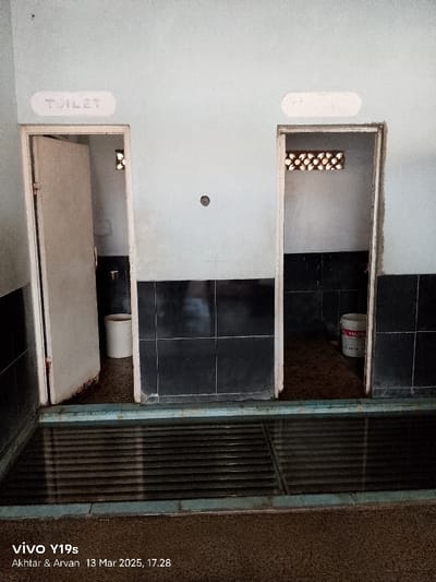 The image shows the interior of a public restroom in Kecamatan Gabuswetan, Indonesia, located at latitude -6.44 and longitude 108.028. The restroom has two stalls, each with a door and a sign above. The sign above the left stall reads "TOILET" and the sign above the right stall is blank. The stalls are separated by a black tiled wall. The floor is tiled in black and gray. There is a white bucket in the right stall, and a white toilet bowl in the left stall. There is a grate on the floor in front of the stalls, and the floor is wet. There are no people in the image. The room is dimly lit and there is a single light fixture visible above the stall doors. The walls are painted a light gray. There is an air vent above the left stall. The image was taken on March 13, 2025 at 5:28 PM.