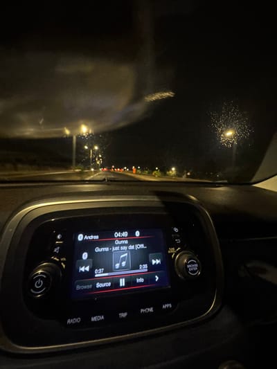 The image captures an interior view from a vehicle at night, looking through a rain-spattered windshield onto a public road. The specific location within Rosh HaAyin, Israel, is not identifiable from the visual evidence.

In the foreground, the upper portion of a vehicle's dashboard is visible, featuring a central infotainment system. The digital display shows "Andrea" connected via Bluetooth, a time of 04:49, and indicates the song "Gunna - just say dat [Offi..." by artist Gunna is playing. The track progress is 0:37 out of 2:35. Playback controls for previous track, pause, and next track are displayed, along with menu options for "Browse," "Source," and "Info." Below the screen are physical buttons labeled "RADIO," "MEDIA," "TRIP," "PHONE," and "APPS," flanked by rotary knobs for power/volume and tune/scroll/enter functions.

Beyond the windshield, a multi-lane road extends into the distance, illuminated by numerous streetlights. Water droplets are visible on the windshield, indicating rain or recent precipitation, causing light reflections. Red tail lights from distant vehicles are discernible along the road ahead against the dark, unlit sky.