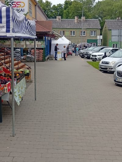 The image shows a street scene in Kuršėnai, Lithuania, likely a market or outdoor event. In the foreground, a stall displays various fruits and vegetables, including watermelon slices, plums, and what appear to be cabbages or lettuces. Several large sacks, possibly containing soil or agricultural products, are stacked behind the stall. To the left, a large blue and white striped awning covers part of the stall area. A yellow building with "SSSS UKAI" and "SENUKAI" signage is visible in the background, suggesting a retail store. Several white tents are set up further down the street. A man in a dark jacket and blue jeans is standing at a counter in one of the tents, interacting with a woman. Other people are seen walking and browsing in the background. A row of parked cars lines the street to the right of the tents. The ground is paved with interlocking gray bricks. The environment is outdoors, with trees visible in the background, indicating a pleasant weather day.