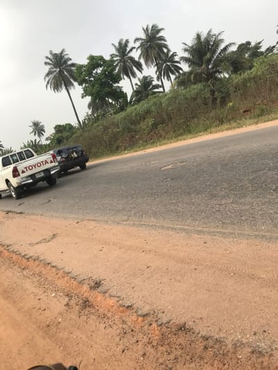 A white Toyota vehicle is parked on the side of an asphalt road, with palm trees lining the opposite edge. The shoulder of the road is dirt and gravel. The sky appears overcast. The overall scene suggests a tropical or subtropical location.