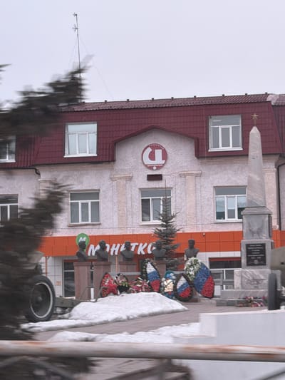 The scene depicts a memorial area in front of a light-colored, multi-story building with a dark red mansard roof, located in Beloyarsky, Russia. The building features white-framed windows, decorative facade elements, and a prominent circular sign on its upper facade displaying "ЦЕНТРАЛЬНЫЙ УНИВЕРСАМ" (Central Department Store/Supermarket). Below this, an orange band runs across the ground floor, bearing a green circular logo with an "M" and the partially visible Cyrillic text "МОНЕТКА" (Monetka supermarket). An antenna structure extends from the roof against an overcast sky.
In the foreground, a war memorial occupies the space. It includes a grey stone obelisk topped with a golden star, positioned to the right. To the left of the obelisk, at least three dark-colored busts of individuals are mounted on pedestals. At the base of the busts and around the memorial, several wreaths are laid, predominantly displaying the red, white, and blue colors of the Russian flag, along with some natural flowers. A small evergreen tree stands among the busts. To the far left, a piece of green artillery, possibly a cannon, with a prominent white-rimmed wheel, is partially visible. Patches of snow cover the ground in the immediate vicinity of the memorial. Blurred foreground elements, including parts of trees and a metal railing, suggest the photograph was taken from a moving vehicle or with camera motion. There are no individuals discernible in the image.