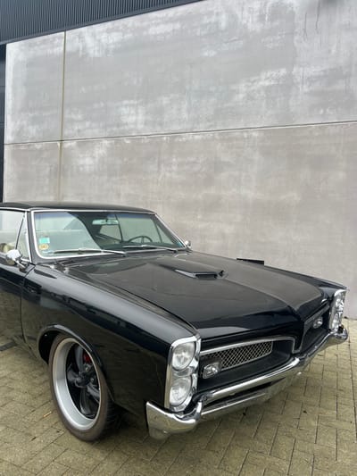 A black, 1960s-era Pontiac GTO hardtop is parked stationary on a light brown herringbone-patterned brick pavement. The vehicle presents a glossy black finish, featuring a prominent chrome front bumper, vertically stacked headlights, a chrome mesh grille, and a raised hood scoop. Custom five-spoke wheels with silver outer rims and visible red brake calipers are fitted to the front axle. A light-colored interior is partially discernible, and a yellow-edged sticker displaying "B E" is affixed to the passenger side of the windshield. The car is positioned in front of a tall, two-tiered, light grey concrete wall showing weathering and discoloration, which is topped by a darker, horizontally ribbed architectural panel. The scene is located in Kortrijk, Belgium. No individuals or active interactions are present.