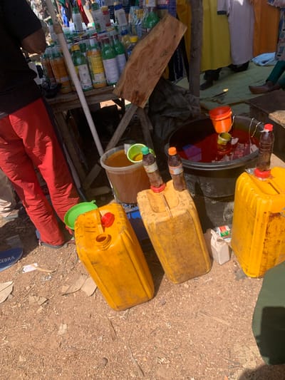 The image depicts an outdoor scene, likely a market stall in Nigeria, showcasing an array of items for sale, primarily liquids in various containers. The setting appears to be dusty and arid, with loose soil and pebbles covering the ground.
Dominating the frame are several large, yellow plastic jerrycans, some filled with a reddish-brown liquid, possibly palm oil or a similar commodity. One jerrycan has a bright red lid and another a green lid. Next to these, a large black basin holds a significant volume of the same reddish-brown liquid, with a few plastic bottles with orange and red caps submerged within it. Smaller plastic bottles, some appearing to contain beverages and others possibly oils, are stacked and displayed on a wooden shelf structure. These bottles vary in color and labeling, suggesting a range of products.
A piece of rough-hewn wood is positioned prominently in the foreground, possibly serving as part of the stall's display or construction. Partially visible in the lower portion of the image are the legs of at least one person, dressed in bright red trousers. Another person's hand, wearing a dark sleeve, is also visible. The overall impression is of a busy market environment where goods are being sold. The lighting suggests it is daytime, with harsh sunlight casting shadows. There is no visible text that is clear enough to be read. The scene conveys a sense of commerce and daily life within a Nigerian context.