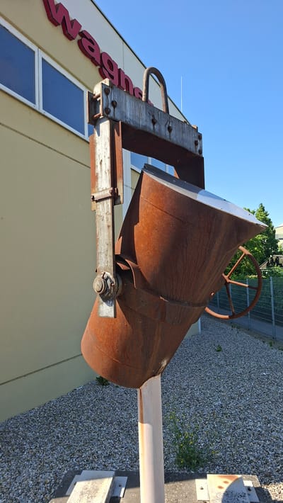 This is a daytime, outdoor, eye-level, medium shot of a rusted metal industrial ladle sculpture displayed in front of a modern building in Enns, Austria. The building, with a light beige facade and two rectangular blue-tinted windows, has a large red sign with white outlines that reads "Wagner" in prominent lettering.

The sculpture itself is a large, rusty ladle with a circular metal wheel attached to its side, suggesting a mechanism for tilting or pouring. The ladle is suspended from a metal frame by what appears to be a heavy-duty hinge and support system. The frame is also made of weathered, rusty metal, bolted together. The ladle is positioned at an angle, appearing to be in a pouring or tipped stance. A white, cylindrical pole supports the ladle and frame structure from below.

The ground surrounding the sculpture is covered in light-colored gravel. In the background, beyond a metal fence, there are trees and a building visible under a clear blue sky. Small green weeds are growing in patches around the base of the sculpture. The lighting suggests a sunny day, with distinct shadows cast by the sculpture. There are no people visible in the image.