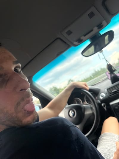 The image depicts the interior of a passenger vehicle, likely a BMW due to the visible logo on the steering wheel, in the city of Krushovitsa, Bulgaria. A male individual is positioned in the foreground, seen from the chest up, with his right profile prominent. He has short dark hair, stubble on his chin, and visible skin discoloration or markings on his right cheek and forehead. His right eye is looking upwards and to the right, and his tongue is partially extended from his open mouth. He is wearing a dark-colored shirt with visible damp patches or stains on the fabric.

The vehicle's driver is partially visible, with their right hand gripping the steering wheel and their left leg, clad in light grey striped shorts, resting in the lower right corner of the frame. The car's interior features include a grey headliner, an overhead console with lights and vents, a rearview mirror from which a dark air freshener or ornament hangs, and a blue tinted strip at the top of the windshield. A black dashboard, an air vent on the passenger side, and keys hanging from the ignition or dashboard are also visible. Through the windshield, an overcast sky with patches of blue is seen above a distant line of green trees and foliage, indicative of a road or highway environment.