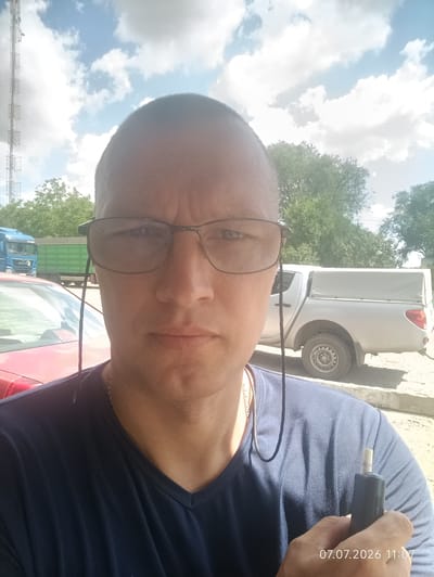 The image is a selfie taken outdoors. The subject, a Caucasian male with a shaved head and wearing glasses and a dark blue V-neck t-shirt, is in the foreground. He is holding a black electronic device with a light-colored tip in his right hand. A thin black wire runs from his ear, down his neck, and is visible against his t-shirt. In the background, there is a red car on the left, and a silver pickup truck with a gray cover over its bed on the right. Behind the vehicles, there are trees and a communication tower. The sky is blue with scattered white clouds. A timestamp in the lower right corner reads "07.07.2026 11:07". The location is identified as Spaske, Ukraine.