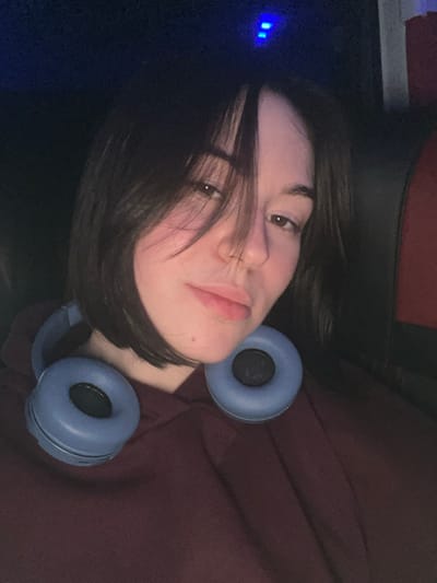 A young person with dark, short hair lies on a dark surface, their face tilted toward the camera. They have fair skin and are wearing light blue headphones. Their hair partially covers their face. A small, mole-like spot is visible on their chin. They wear a reddish-brown garment, possibly a sweatshirt. Two blue lights are visible in the background on the left side of the image.