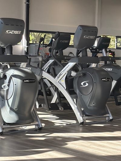The image shows several Cybex Arc Trainers in a gym. The machines are gray with silver frames. The machines are angled towards the right side of the frame, with the rightmost machine casting shadows on the floor. Windows can be seen in the background.