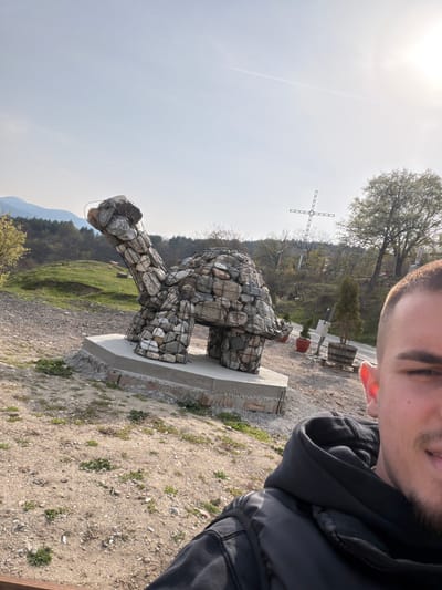 A selfie captured in the area of Sveti Konstantin, Bulgaria, shows a man with short hair and a beard wearing a dark hooded jacket, positioned in the upper right of the frame. A large, artistic stone turtle sculpture stands on a concrete base in the center of the image. In the background, a metal cross is visible on a hill, surrounded by trees and vegetation. The environment consists of a gravel area with sparse plant growth. Mountains are visible in the distance under a bright sky.
