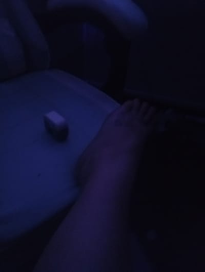 The image depicts a low-angle, dimly lit interior scene, likely a bedroom or living area, with a strong blue-purple ambient light. A person's bare foot, with visible toes, is positioned in the lower right quadrant of the frame, resting on a soft, possibly upholstered surface. The foot appears to have a faint tattoo on the ankle area. To the left of the foot, on the same surface, is a small, rectangular object, possibly an eraser or a small electronic device, its details obscured by the low light. The background is dark and indistinct, with suggestions of furniture or architectural elements. There are no discernible features suggesting the city of Viviers, France, or any specific geographic location.