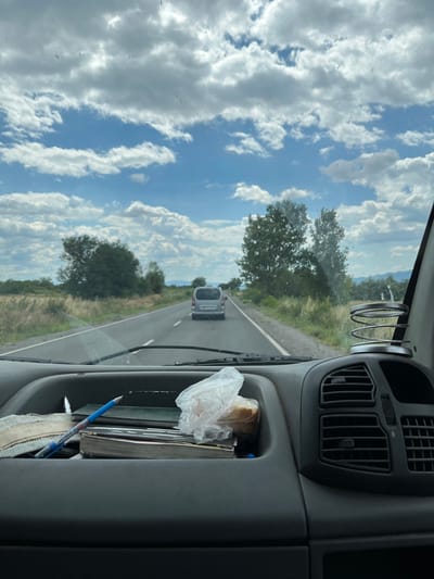 The image shows a view from inside a vehicle traveling on a road. The road has a white line marking the right lane and a double yellow line on the left. A silver minivan is visible ahead on the road. The landscape on either side of the road consists of dry grass and scattered trees. The sky is filled with white and grey clouds against a blue backdrop. In the foreground, the dashboard of the vehicle is visible, with a compartment holding several items, including books, pens, and a plastic bag containing bread. A spring-loaded cup holder is also present on the right side of the dashboard. The context of the location being in the city of Pevtsite, Bulgaria, is provided externally and is not visually discernible from the image itself.