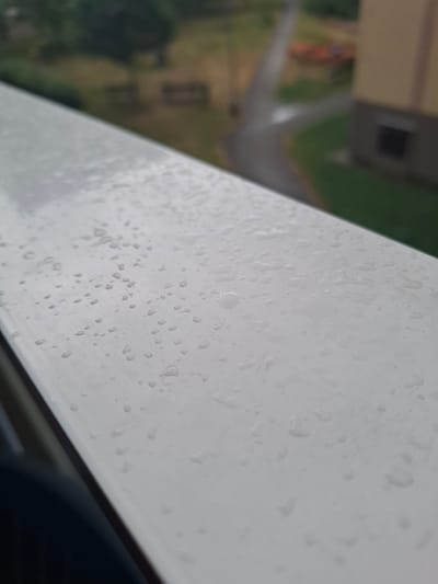 The image shows a close-up of a wet, white railing with numerous water droplets on its surface. The railing is positioned diagonally, occupying the majority of the foreground. In the blurred background, a paved path winds through a grassy area with some trees and a building. The building appears to be light-colored with a dark rectangular window. The scene suggests it is raining or has recently rained. The location is Ústí nad Orlicí, Czechia.