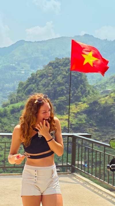 A woman with curly red hair is laughing with her hand over her mouth on a balcony. She is wearing a black crop top, white shorts and sunglasses on her head. She is holding a small object in her other hand. In the background there is a Vietnamese flag waving in the wind against a backdrop of green mountains. There is a metal railing separating the balcony from the mountains. The scene is set in Lũng Phìn Commune, Vietnam.