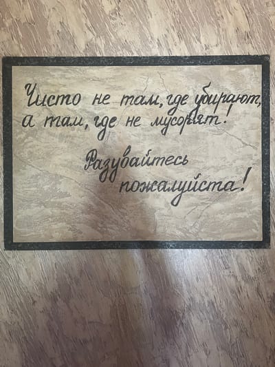 A black-bordered rectangular sign with a textured, marble-like background is affixed to a wall with a wood-grain pattern. The sign displays a handwritten message in Cyrillic script. The text translates to "It's not clean where they clean, but where they don't litter. Please don't litter!". The sign is located in an indoor environment, possibly a public restroom or common area, in Bolshoy Molokish, Moldova. There are no people or animals visible in the image.