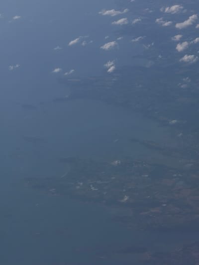 This is an aerial photograph of a coastal area, likely taken from an airplane. The image captures a vast expanse of blue water, dotted with scattered white clouds. The lower portion of the image shows a hazy view of land, with hints of vegetation and some geometric patterns that could indicate urban development or agricultural fields. A faint, curved line suggests a coastline. There are no discernible people, specific objects, or actions depicted in the image, other than the natural elements of water, clouds, and land. The overall impression is one of an expansive, serene landscape viewed from a high altitude. The location is described as Unknown, France, but without specific landmarks, pinpointing a precise location within France is impossible.