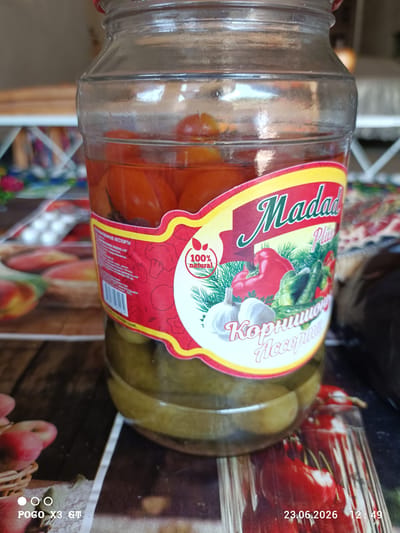 This is a medium shot of a glass jar containing pickled vegetables and tomatoes, sitting on a patterned surface. The jar has a red lid and a prominent label with a yellow and red border. The label displays the brand name "Madad Plus" in a stylized font, along with an illustration of vegetables like bell peppers, cucumbers, garlic, and dill. Text on the label is in Cyrillic, indicating "КОНЫРОВАННЫЕ АССТОРТИ" (Marinated Assortment) and "Корнишон Ассорти" (Gherkin Assortment). There's also a "100% NATURAL" seal on the label.
The background is blurred, but hints of furniture and possibly a window suggest an indoor setting. The surface the jar is on appears to be a table with a wood-grain pattern and colorful illustrations of fruits like apples and cherries. In the bottom left corner of the image, there is text indicating a phone model "POGO X3 GT." In the bottom right corner, a date and time stamp reads "23.06.2026 12:49". The lighting suggests it is daytime. The overall impression is a still life arrangement showcasing a food product.