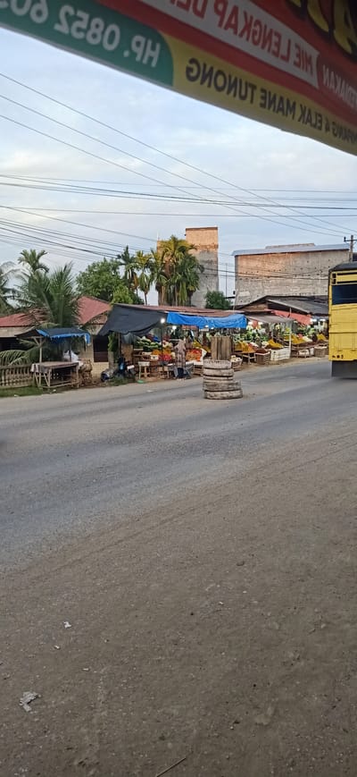This image depicts a roadside market scene in Gampongteungoh, Indonesia, likely in the late afternoon. The sky is overcast with diffused light, suggesting a cloudy day or approaching evening. The foreground is dominated by a paved road, with a granular texture and scattered debris.
To the left of the road, a row of market stalls is visible. These stalls are covered with various tarps, including black and blue ones, providing shade for the produce displayed. Various fruits, such as watermelons and what appear to be pineapples, are arranged on wooden tables and crates. A person wearing a headscarf and traditional clothing is standing behind one of the stalls, seemingly attending to customers or organizing the goods. A motorcycle is parked near the stalls, indicating local transport. A small building with a red tiled roof is situated behind the initial stalls.
Further down the road, more market stalls extend, displaying a wide assortment of fruits and vegetables. The stalls are simple wooden structures, some with corrugated metal roofing. In the middle of the road, a stack of used tires serves as a visual marker or barrier. To the far right, the rear of a yellow vehicle, possibly a truck, is partially visible, suggesting ongoing traffic.
In the background, several buildings are present. One is a multi-story structure with a flat roof, appearing to be under construction or an industrial building. Another is a long, low building with a lighter colored roof. Palm trees and other tropical foliage are scattered throughout the scene, reinforcing the Indonesian location. The overall atmosphere is that of a bustling, everyday market, with a focus on fresh produce.
A prominent sign is visible at the top of the image, reflected or attached to an overhead structure. The text on the sign is mostly mirrored and partially obscured, but some legible parts include "HP .0825", ".9H", and what appears to be Indonesian words like "LENGKAP" and "MINUMAN". This likely indicates a business or advertisement related to a phone number or a shop selling beverages or other goods.