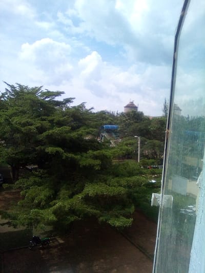 The image depicts an outdoor scene observed from an elevated position through a partially open window in West Sakwa ward, Kenya. The foreground on the right features a metallic window frame with a glass pane, reflecting distorted portions of the interior and exterior; the adjacent wall is painted a light blue-grey, showing areas of peeling paint.
The dominant mid-ground and lower left are characterized by dense, lush green tree canopies. Beneath the trees in the lower left, a black motorcycle is parked on a dark ground surface, with a light-colored vehicle partially visible further left. Amidst the trees in the mid-ground, an elevated structure with a distinctive blue roof and visible stairs is present, alongside a tall light pole. Above the tree line in the distant background, two prominent, light-colored, cylindrical structures, each topped with a dark, conical roof, are visible, suggestive of water towers or similar utility buildings. The sky above is partly cloudy, displaying significant white cumulus clouds against a clear blue expanse. No individuals are visible within the frame.