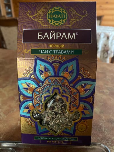 The image displays a close-up of a box of HAYATI brand tea. The box has a predominantly purple and gold design with intricate mandala-like patterns in shades of blue, orange, and teal. The front of the box features text in Russian. Prominently displayed is the word "БАЙРАМ" in large white letters within a dark maroon rectangle, indicating the product's name. Above this, in smaller yellow text, is "ЧЁРНЫЙ" (black), and below it, in green text, is "ЧАЙ С ТРАВАМИ" (tea with herbs). A cut-out window on the box reveals the contents: a mixture of dried herbs and tea leaves. Towards the bottom of the box, more Russian text reads "ЧАЙНАЯ КОЛЛЕКЦИЯ ТАТАРСТАНА" (Tea Collection of Tatarstan) and "ВЕС НЕТТО: 70г" (Net Weight: 70g). There is also a small circular logo with a stylized design and what appears to be text within it. The box is placed on a brown, textured surface, possibly a table, in an indoor setting. The background is blurred, showing hints of furniture and possibly wood paneling. The lighting suggests an indoor environment, with no specific identifiable landmarks of Dimitrovgrad, Russia.
