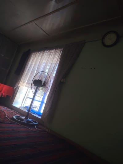 The image depicts an interior scene, possibly a room in a dwelling. A floor fan is positioned in front of a window that is covered by sheer, patterned curtains. The window appears to be open, allowing natural light to filter through, with a blue tint visible from the outside. On the floor, a carpet with a striped pattern of red and dark grey covers the surface. To the left of the fan, a table covered with a red cloth is partially visible. On the wall to the right, a round clock with a dark frame is mounted, showing the time. The walls are painted a light green color. There are three small dark dots arranged horizontally on the wall above the clock. The ceiling appears to be made of panels, with some discoloration and damage visible. The overall lighting is dim, with strong contrast between the illuminated window area and the shadowed parts of the room. The image is taken from a low angle, looking upwards towards the ceiling. The location is specified as Almachi, Uzbekistan.