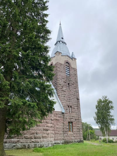 An exterior, eye-level, full shot captures the stone tower of a church in Melnikovo, Russia, on an overcast day. In the foreground to the left, the dense branches of a large evergreen tree obscure a portion of the church's facade. The tower is constructed of rough-hewn, reddish-brown stones, with a sloped, gray metal roof topped by a pointed spire. A tall, slender birch tree stands to the right of the tower. Beyond the church and trees, a dirt road leads past a few buildings with gabled roofs. The sky is a uniform, pale gray, indicative of cloudy weather.