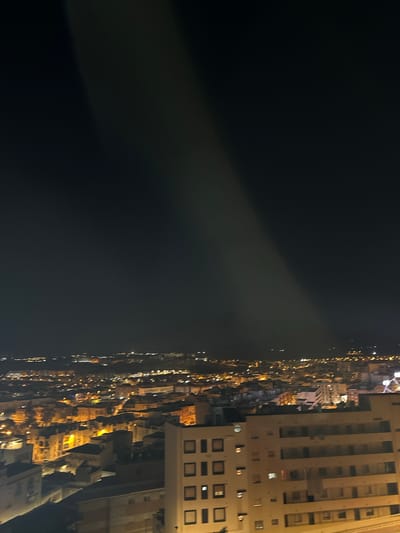 A nighttime aerial view of Puerto De Mazarrón, Spain. The foreground features a light-colored, multi-story building with numerous windows, some illuminated. Beyond this building, the city stretches out, characterized by a dense collection of residential and commercial structures, many displaying illuminated windows in warm orange and yellow hues. The distant landscape is dotted with more lights, suggesting the expanse of the city and its surroundings. A faint, diagonal beam of light cuts across the dark night sky, originating from the upper left quadrant and extending downwards towards the cityscape. The overall scene depicts an urban environment at night, bathed in artificial light.