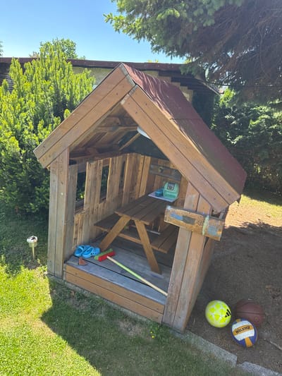 A slightly high-angle shot displays a children's playhouse with a reddish-brown roof and wooden construction. The playhouse is set in a garden or yard, with grass and foliage visible around its base. Inside, a child-sized table and items are seen, including blue sandals and a broom. Next to the playhouse are three balls. The photo was taken during daylight. There is no evidence to indicate that the image was taken in the city of Zürchau, Germany.