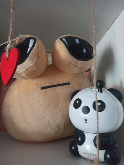 The image displays a close-up view of two stuffed toys, likely on a shelf. On the left is a large, light brown, vaguely animal-shaped plush toy with two large black eyes outlined in white, and a small red heart hanging from a string on its left side. A thin black horizontal line is visible on its body. To the right of this larger toy is a smaller, white and black stuffed panda bear. The panda has black ears, black eye patches, and a black mouth. It appears to be a pencil sharpener, with a circular opening on its forehead. A piece of twine runs vertically through the center of the image, obscuring part of the panda. The background is a pale, greyish surface, possibly a wall or the interior of a shelf. The lighting suggests an indoor environment. There is no visible indication of the city of Promyshlennaya, Russia, within the image itself.