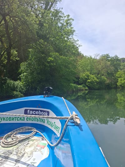 The image shows the bow of a bright blue boat on a body of water with green foliage and trees visible in the background. The water's surface reflects the surrounding trees and sky. A rope is tied to the boat's cleat. A sticker on the boat features the Facebook logo and Bulgarian text. Based on the text on the sticker and likely geographic location, the image is potentially taken in the region of Petrevene, Bulgaria.
