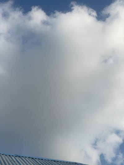 The image depicts a view of a cloudy sky with a sliver of a building on the right edge. The sky is filled with large, diffused white clouds that dominate the frame, obscuring much of the blue sky beneath. The clouds appear soft and voluminous, suggesting a partly cloudy or overcast day.

On the far right, a portion of a modern building is visible. Its exterior is characterized by corrugated metal panels, arranged in a vertical pattern with distinct ridges and valleys. These panels are a neutral gray color, and the metallic texture is evident. Small rivets or fasteners can be seen along the seams of the panels, indicating construction details. The building's angle suggests it is tall and may be a contemporary structure.

There is no discernible activity or event taking place; the image is a static shot of the sky and a building. The time of day is not explicitly indicated, but the bright, diffused light from the clouds suggests it is daytime, possibly mid-morning or afternoon. The weather appears to be cloudy, potentially with some sun breaking through in places, given the brightness of the clouds. The location is identified as Pantinapu, Nigeria, but there are no specific visual cues in the image to confirm this location beyond the given context. No people or text are visible in the image. The overall mood conveyed is one of a typical daytime sky, perhaps with a slight sense of awe at the scale of the clouds and the geometry of the building.