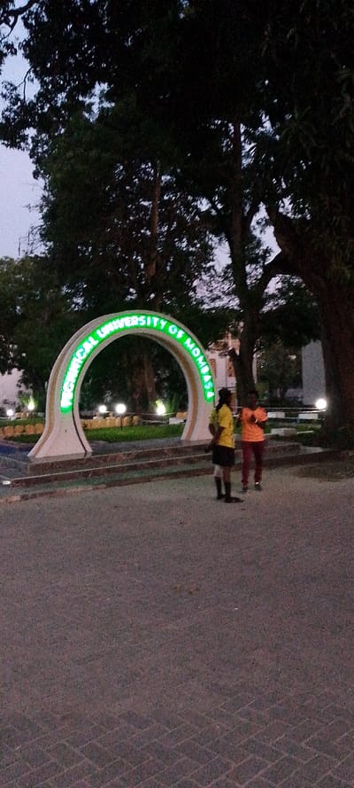 The image depicts an outdoor scene at dusk or night, located at the Technical University of Mombasa in Tudor ward, Kenya. A prominent white arched monument stands centrally, featuring the words "TECHNICAL UNIVERSITY OF MOMBASA" illuminated in bright green neon or LED lights along its inner curve. The monument is set on a stepped base, with several spherical, ground-level lights illuminating a surrounding grass patch.
Two individuals are visible in the foreground, positioned to the right of the monument. The person on the left wears a yellow t-shirt and dark shorts or skirt, with dark footwear, facing towards the right. The person on the right wears an orange t-shirt and dark trousers, also with dark footwear, facing towards the left, positioned opposite the first individual. The ground is paved with dark interlocking blocks laid in a herringbone pattern.
In the background, numerous large, dark trees with dense foliage are silhouetted against a fading twilight sky, which transitions from light blue near the horizon to darker tones overhead. Faint outlines of buildings are discernible through the trees on the right side of the frame.