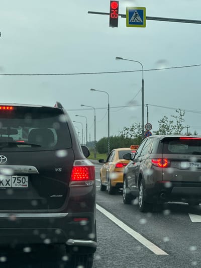 In Раздолье Снт, Russia, on a cloudy day, traffic is stopped at a red light. The foreground shows the rear of a dark-colored SUV. To its right, a brown SUV is visible with its rear lights illuminated. Further ahead, a yellow sedan, likely a taxi, is stopped. Overhead, a traffic light displays a red signal and a countdown timer showing "26". Adjacent to the traffic light is a pedestrian crossing sign. Several streetlights are visible along the road, and trees line the background. The road surface appears wet, suggesting recent rain.