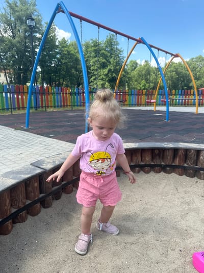 A young, blonde-haired girl with her hair in a bun stands in a sandbox in an outdoor playground in Donchovci, Bulgaria. She is wearing a pink t-shirt with a cartoon character on it and pink shorts. Her shoes are also pink. The sandbox is surrounded by a wooden border. In the background, there is a colorful fence, a swing set, and trees. The sky is blue with some clouds. The ground of the playground is covered with black and red rubber tiles.