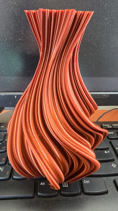 An orange, 3D-printed vase with a twisted, ribbed design rests on a black laptop keyboard. The vase is centrally located in the frame, with its widest point towards the bottom and tapering towards the top, where the ribs are more tightly packed and appear to flare slightly outwards. The keyboard is visible beneath the vase, with some keys such as "F6," "Alt," and "Ctrl" discernible. The background is dark and textured, possibly a countertop or a dark surface. The lighting appears to be from above and slightly to the side, casting subtle shadows on the vase and the keyboard. There are no people or any indication of specific location within Krasnopol'ye, Russia.