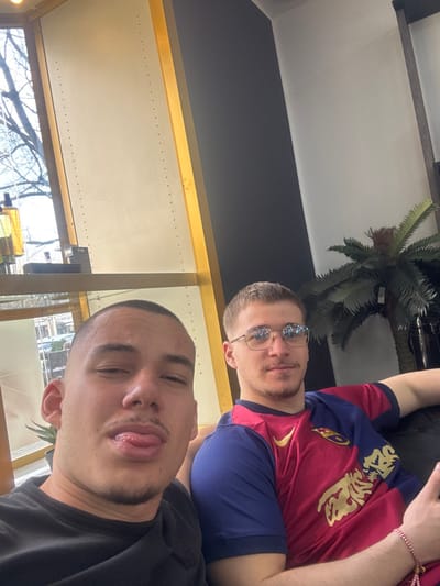 This image depicts two young adult males indoors in what appears to be a modern commercial establishment in Krems an der Donau, Austria.
The foreground subject, positioned to the left, is a male with a closely-cropped buzz cut and light stubble. He wears a black short-sleeved t-shirt and is making a playful expression, sticking his tongue out slightly while looking directly at the camera.
To his right and slightly behind him, a second young adult male is seated on a dark-colored, upholstered surface, possibly a couch or armchair. He has short, light brown hair, a short beard, and wears rectangular-framed eyeglasses. He is dressed in a red and blue short-sleeved soccer jersey (resembling an FC Barcelona kit) and has a multi-colored beaded bracelet on his left wrist. This individual is looking towards the camera with a subtle smile. His left arm is casually resting on the back of the seat.
The background includes a large window to the far left, revealing outdoor elements such as bare tree branches and some distant structures, suggesting an urban street view. A horizontal shelf or counter runs beneath the window, holding a dark object and a bottle containing yellow liquid. A prominent vertical yellow-gold beam or paneling borders the window structure. The wall behind the seated male is dark grey, transitioning to a lighter color higher up. An artificial palm-like plant in a dark pot is visible in the upper right corner of the frame. The scene is brightly lit, with natural light entering from the window.