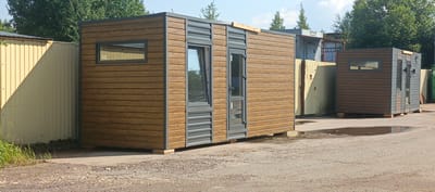 The image shows a small, prefabricated modular building with a wood-like exterior siding and gray trim, parked on a gravel lot. The building has one rectangular window and a glass-paneled door with gray trim. To the left of the building is a tall, corrugated metal fence, painted light yellow. In the background, there are trees, other buildings, and what appear to be industrial structures. The scene is set in an outdoor industrial or storage yard. The ground is covered with gravel and there are a few small puddles of water. The sky is blue with some clouds. The building appears to be in a lot located in Sosnoviy Bor, Russia, a city known for its industrial areas and proximity to the Baltic Sea.