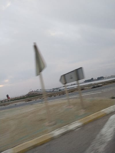 A blurry roadside view under a cloudy sky. Two road signs, a triangular one and a rectangular one, stand beside a highway. Vegetation lines the median. The road stretches towards a distant horizon, where buildings and possibly water are visible. A red stop sign is also visible on the left. The overall blur suggests motion or a shaky camera. The color palette is muted, dominated by grays and browns.