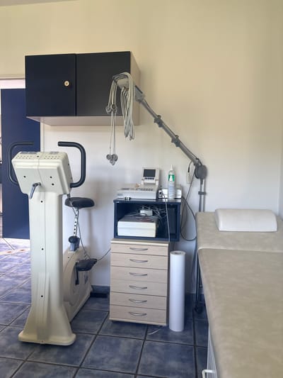 In a room with tiled floors and plain walls, a medical or rehabilitation setting is depicted. Dominating the left side is a cream-colored stationary bicycle with black handlebars and a seat. To the right of the bicycle, a dark blue cabinet with two doors hangs on the wall, containing what appears to be medical equipment and wires. Below this cabinet is a light wood chest of drawers with silver handles, and to its right, a roll of white paper. A grey adjustable arm with wires and sensors is attached to the wall above the cabinet. Further to the right, a medical examination table with a white headrest is positioned. The overall impression is of a clinical or therapeutic space.