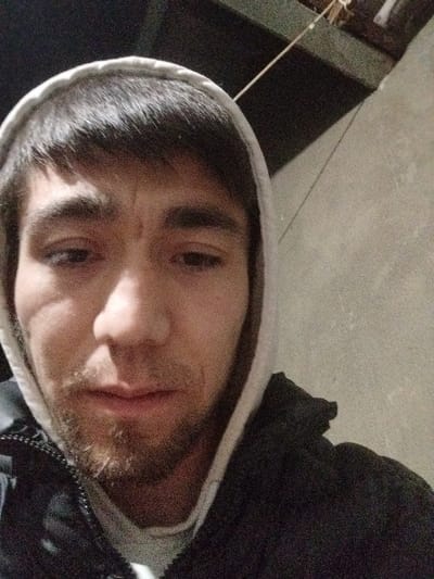 A close-up selfie reveals a man with dark hair and a light-colored hooded sweatshirt. He has a stubbly beard and appears to be in his late twenties or early thirties. A scar is visible near his left eye. The background is a dark, possibly indoor, setting with a rough, textured wall. He is wearing a dark jacket under the hoodie. The lighting is dim, casting shadows on his face. His expression is neutral.
