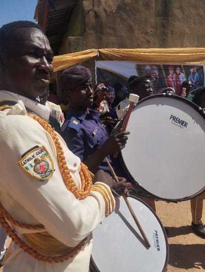 Two men stand beside drums. The first man wears a white uniform with gold detailing and a Catholic Cadet patch. The second man wears a navy uniform and beret, holding a mallet over a large drum. Both appear to be part of a procession or performance. Drums are clearly labelled "Premier England". A banner hangs in the background, showing several people. The building structure is behind the banner and the sky is visible at the top of the frame.