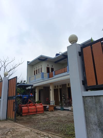 An exterior forensic image depicts a two-story residential property located in Clacuc, Timor-Leste, viewed from a slightly low angle under an overcast sky. The house, positioned centrally, features a light cream-colored facade with blue trim accents on its eaves and balconies. The first floor includes multiple windows and at least one visible wooden door, with a stone-clad section on the exterior wall. A balcony on the second floor presents a light blue patterned railing and reddish-brown, wood-toned elements. A blue water tank and an antenna are visible on the roofline.
To the left of the house, a blue tractor, equipped with a large red rotary tiller attachment, is partially parked behind an open gate. The gate comprises light grey concrete posts, one on the right with a white spherical light fixture, and brown, wood-textured panels. The ground in the foreground is unpaved, consisting of dirt, gravel, and sparse vegetation, with a section of concrete paving directly in front of the house. A bare-branched tree is visible behind the left gate post. On the paved area near the house, a rustic wooden bench or table and several chairs are present, along with potted plants. No individuals are visible in the scene.