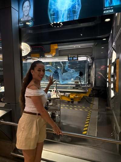 An adult female stands smiling and gesturing within an indoor, artificially lit, themed environment at Pandora – The World of Avatar in Disney's Animal Kingdom, Lake Buena Vista, Florida, near Kissimmee, United States.
The female subject has long, dark, wavy hair and wears a white off-the-shoulder top, light brown pleated shorts, and a dark brown belt. She is positioned with her right arm raised, hand open and fingers splayed, pointing towards a central transparent, cylindrical containment unit. This unit is filled with liquid and contains a light blue, humanoid figure, characteristic of a Na'vi avatar, suspended in a prone or fetal position.
Above the containment unit, a large digital display screen shows a blue, X-ray-like image of a torso and a portrait of a woman labeled "RIVER" with an adjacent "9." Text on the screen also reads "CLONE BATCH 32-C AVATAR." The background features industrial-style yellow and grey structures with exposed piping. To the left of the subject, a metal utility cart holds laboratory equipment, including beakers and vials. The floor is grey with yellow and black striped hazard markings and drainage grates. A metal handrail extends across the lower foreground. A partial reflection of an individual wearing a red baseball cap is visible in a glass surface on the far right.